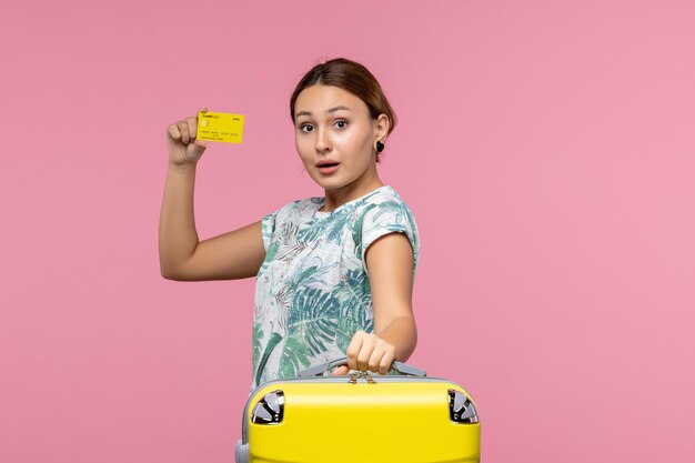 분홍색 벽에 노란색 은행 카드와 휴가 가방을 든 젊은 여성의 전면