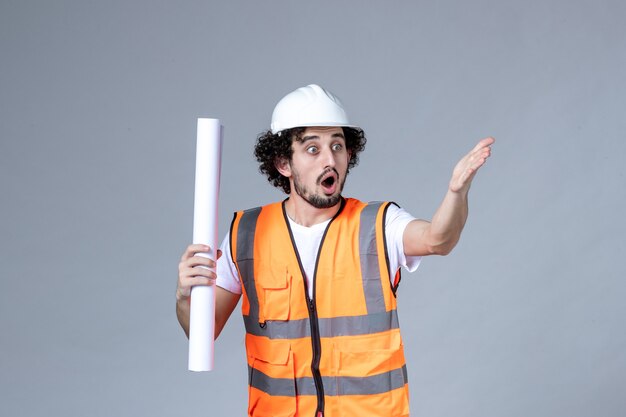 Front view of young wondering male constructor in warning vest with safety helmet and showing blank on gray wall