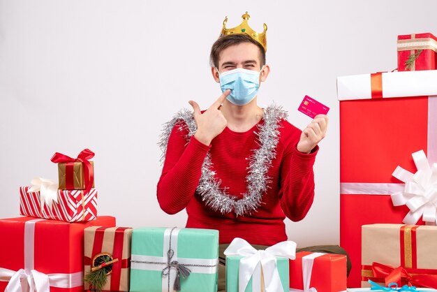Вид спереди молодой рождественский человек с маской, держащий карточку, сидя на полу рождественские подарки