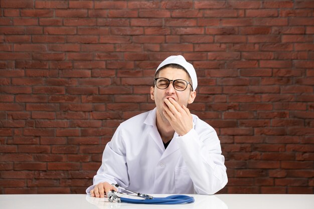 Front view young yawning doctor in white medical suit on brown brick wall