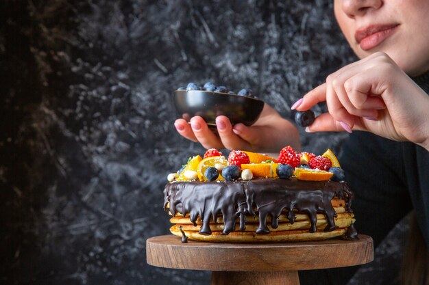 Front view yummy chocolate cake getting decorated with fruits by female on dark wall