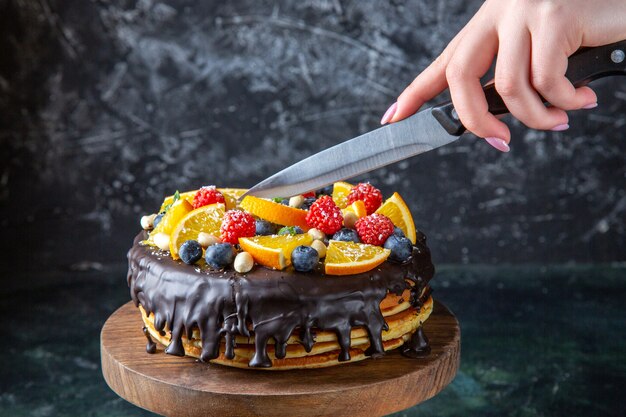 Front view yummy chocolate cake with fruits getting cut by female on dark wall