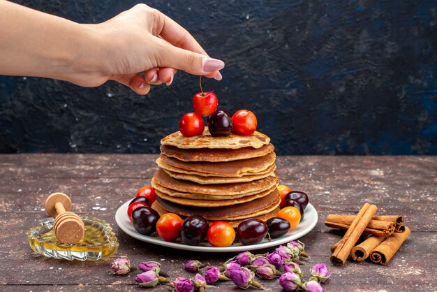 A front view yummy pancakes with cherries inside white plate with flowers and cinnamon on the dark background fruit pancake 