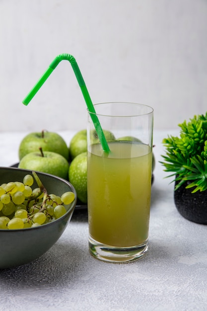 Frontal view apple juice in a glass with a green straw  with green grapes and green apples in bowls on a white background
