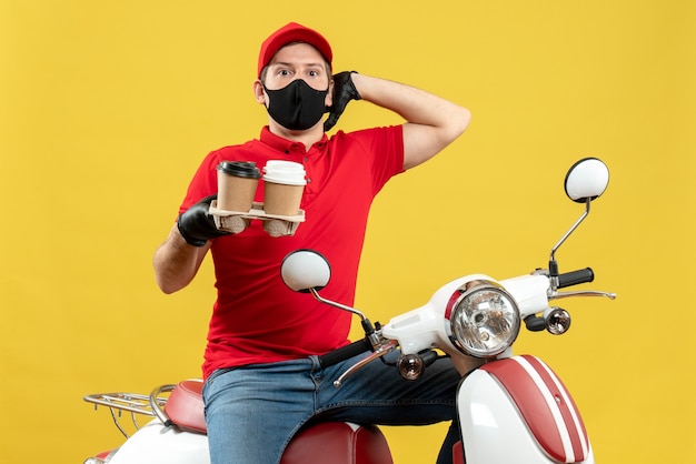 Frontview of surprised delivery guy wearing uniform and hat gloves in medical mask sitting on scooter showing orders