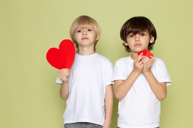 A frotn view two boys in white t-shirts and jeans holding heart shapes on the stone colored space