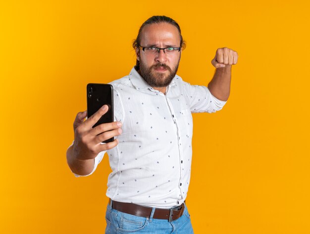 Frowning adult handsome man wearing glasses stretching out mobile phone towards camera showing fist 