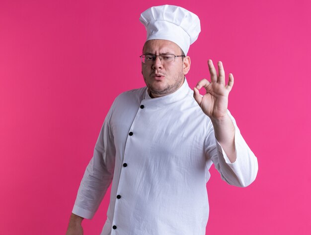 frowning adult male cook wearing chef uniform and glasses standing in profile view looking at front doing ok sign isolated on pink wall