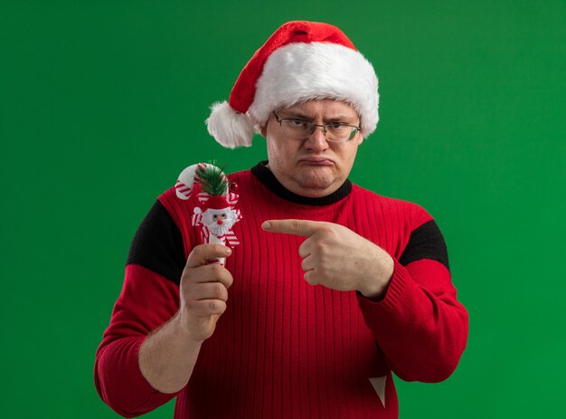 Frowning adult man wearing glasses and santa hat holding pointing at candy cane ornament looking at camera isolated on green background