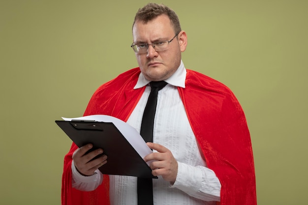 Frowning adult superhero man in red cape wearing glasses and tie holding clipboard looking at front isolated on olive green wall