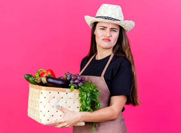 Foto gratuita aggrottando le sopracciglia bella ragazza giardiniere in uniforme che indossa cappello da giardinaggio tenendo il cesto di verdure isolato sul colore rosa