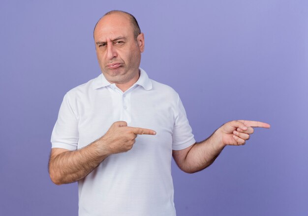 Frowning casual mature businessman looking at camera pointing at side isolated on purple background