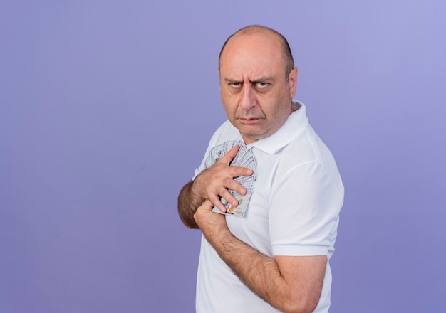 Frowning casual mature businessman standing in profile view holding money isolated on purple background with copy space