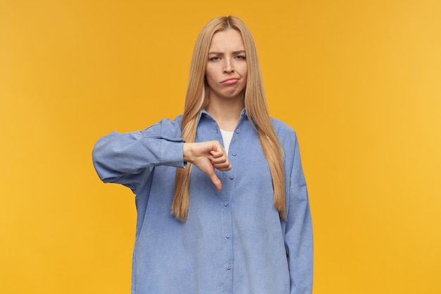 Frowning girl, discontent looking woman with blond long hair. wearing blue shirt. people and emotion concept. showing thumb down, disapproval. watching at the camera, isolated over orange background