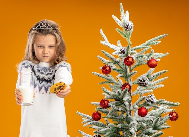 Бесплатное фото Хмурится маленькая девочка, стоящая рядом с елкой в тиаре с гирляндой на шее, протягивая стакан молока с печеньем перед камерой, изолированной на оранжевом фоне