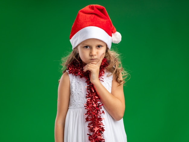 Free photo frowning little girl wearing christmas hat with garland on neck putting hand on chin isolated on green wall