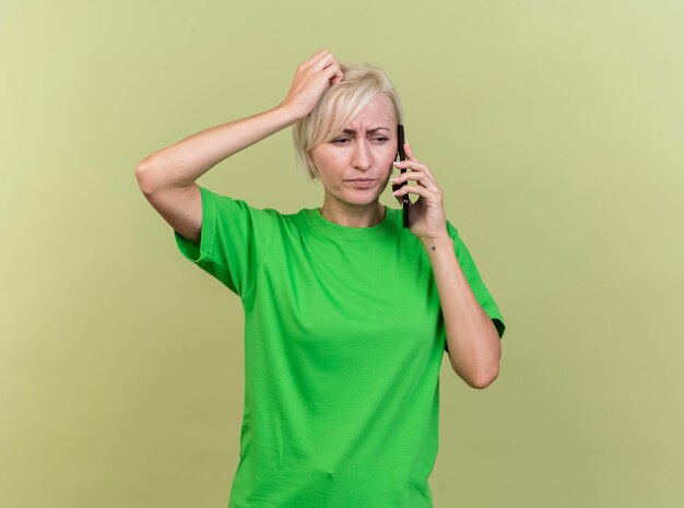 Frowning middle-aged blonde slavic woman talking on phone looking at side putting hand on head isolated on olive green wall with copy space