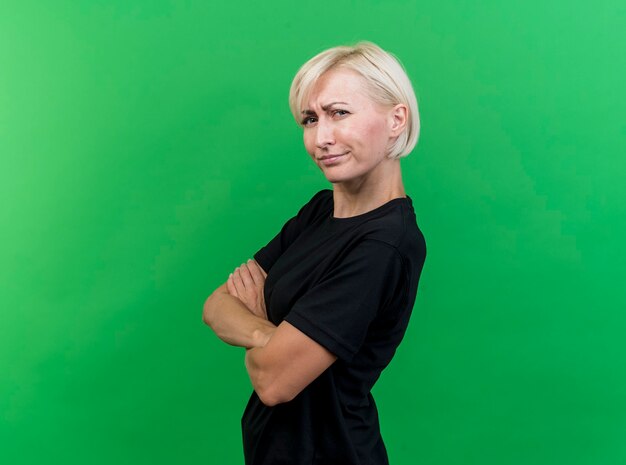 Frowning middle-aged blonde woman standing with closed posture in profile view looking at front isolated on green wall