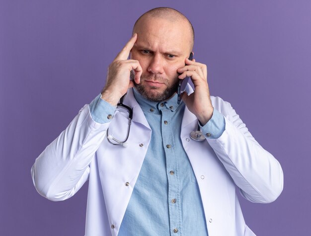 Frowning middle-aged male doctor wearing medical robe and stethoscope talking on phone looking down doing think gesture isolated on purple wall