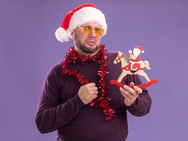 Frowning middle-aged man wearing santa hat and tinsel garland around neck with glasses holding santa on rocking horse figurine  isolated on purple wall