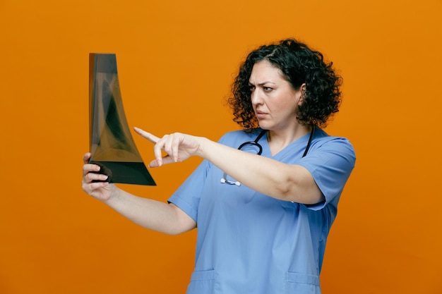 хмурая женщина-врач средних лет в униформе и со стетоскопом на шее, стоящая в профиль, держащая рентгеновский снимок, глядя на него и указывая на него изолированно на оранжевом фоне