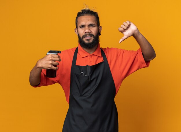 Нахмурившийся молодой афро-американский парикмахер в униформе держит лак для волос, показывая большой палец вниз на оранжевой стене