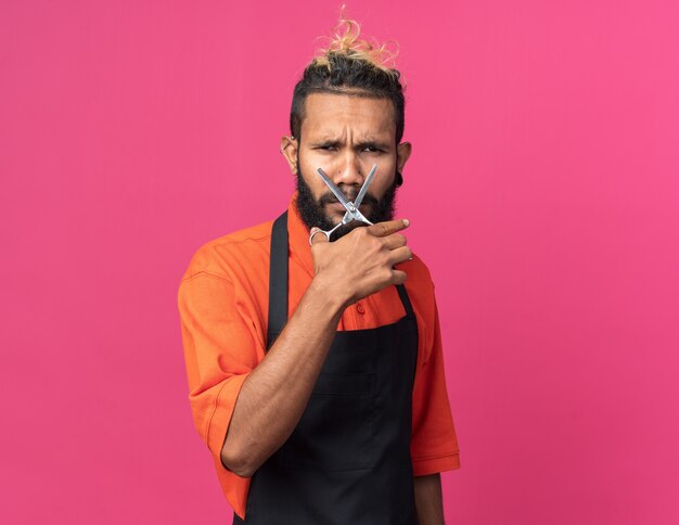 Frowning young afro-american male barber wearing uniform holding scissors in front of face 