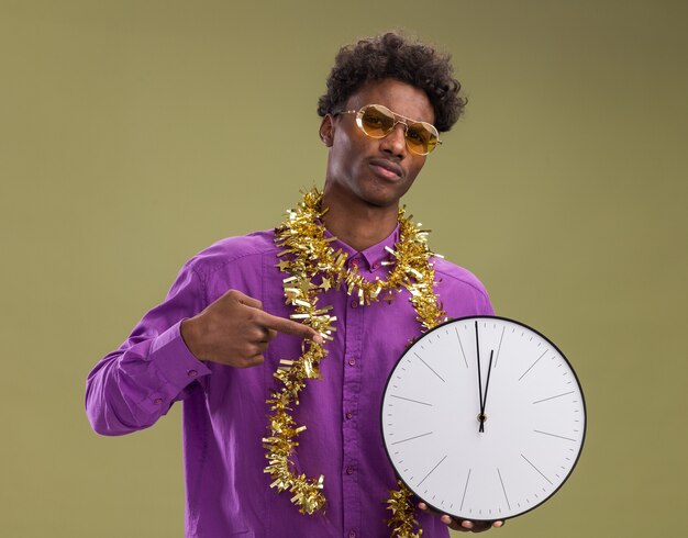 Хмурый молодой афро-американский мужчина в очках с гирляндой из мишуры на шее держит и указывает на часы, глядя в камеру, изолированную на оливково-зеленом фоне