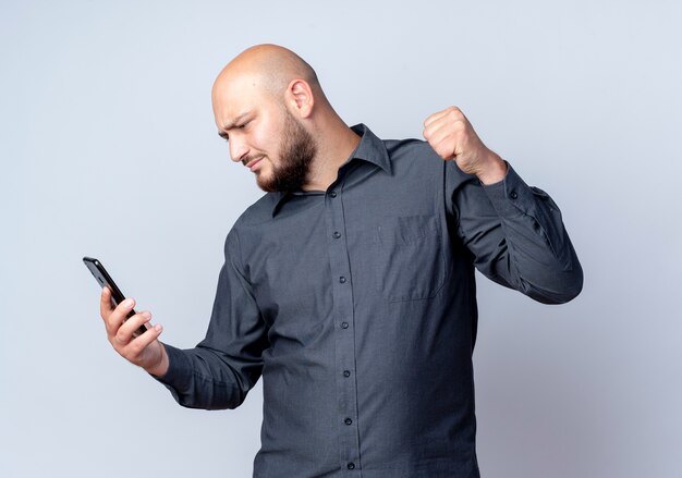 Frowning young bald call center man holding and looking at mobile phone and raising fist isolated on white 