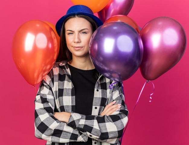 Free photo frowning young beautiful girl wearing party hat standing in front balloons crossing hands