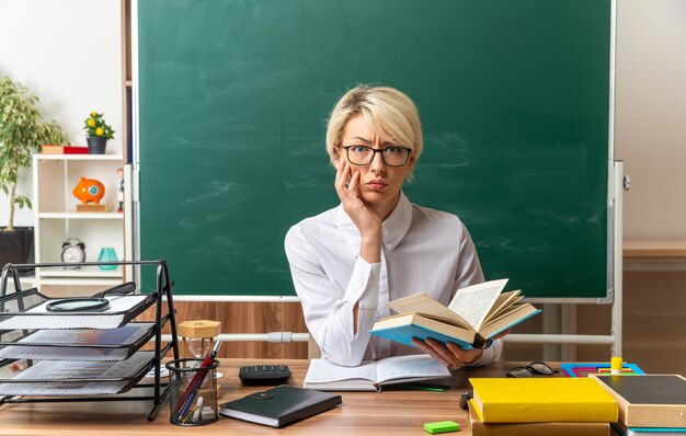Нахмурившись, молодая блондинка учительница в очках сидит за столом со школьными инструментами в классе, держа открытую книгу, держа руку на лице, глядя в камеру