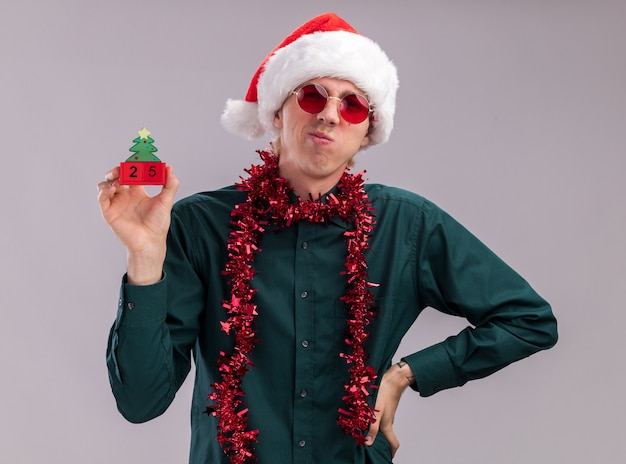 흰색 배경에 고립 된 허리에 손을 유지 카메라를보고 날짜와 크리스마스 트리 장난감을 들고 목 주위에 반짝이 갈 랜드와 산타 모자와 안경을 쓰고 찡그림 젊은 금발의 남자