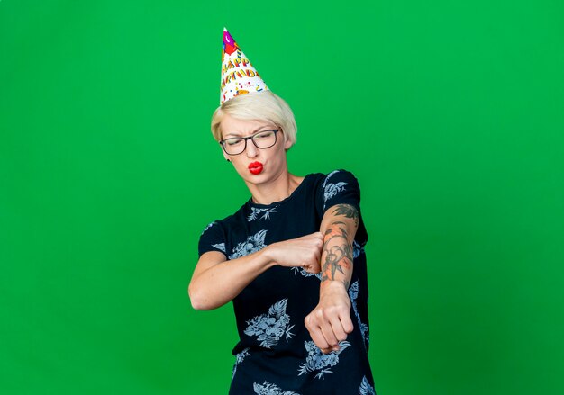 Frowning young blonde party girl wearing glasses and birthday cap stretching out fist touching arm with fist looking at arm isolated on green background with copy space