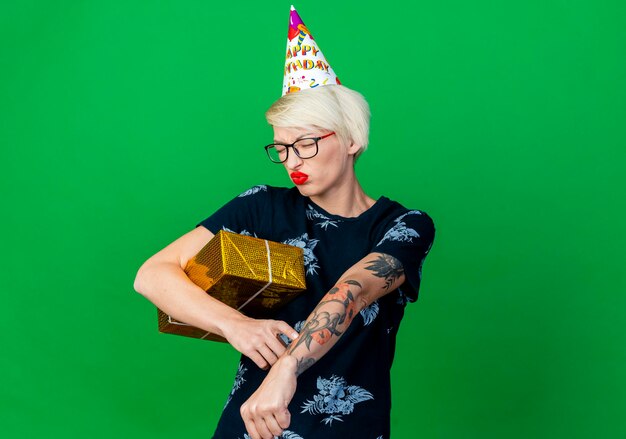 Frowning young blonde party woman wearing glasses and birthday cap holding gift box doing you are late gesture with closed eyes isolated on green wall with copy space