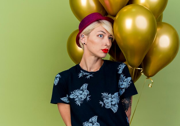 Frowning young blonde party woman wearing party hat standing in front of balloons looking at side keeping hands behind back isolated on olive green wall with copy space