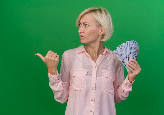 Free photo frowning young blonde slavic woman holding money turning head to side and pointing at side isolated on green background with copy space