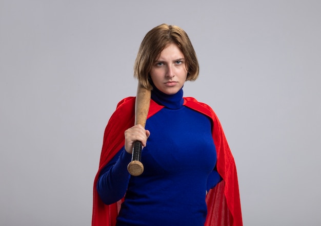 Frowning young blonde superwoman in red cape holding baseball bat on shoulder looking at front isolated on white wall