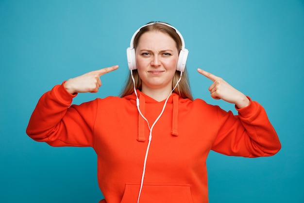 Frowning young blonde woman wearing headphones pointing at them listening to music 