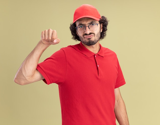 Frowning young caucasian delivery man in red uniform and cap wearing glasses  doing knocking gesture isolated on olive green wall