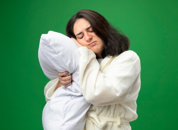 Frowning young caucasian ill girl wearing robe standing in profile view hugging pillow putting head on it isolated on green background