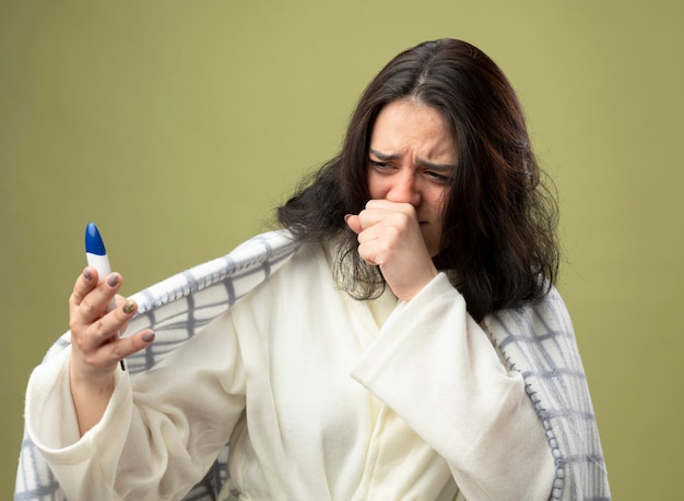 Frowning young caucasian ill girl wearing robe wrapped in plaid holding and looking at thermometer coughing keeping fist near mouth isolated on olive green background