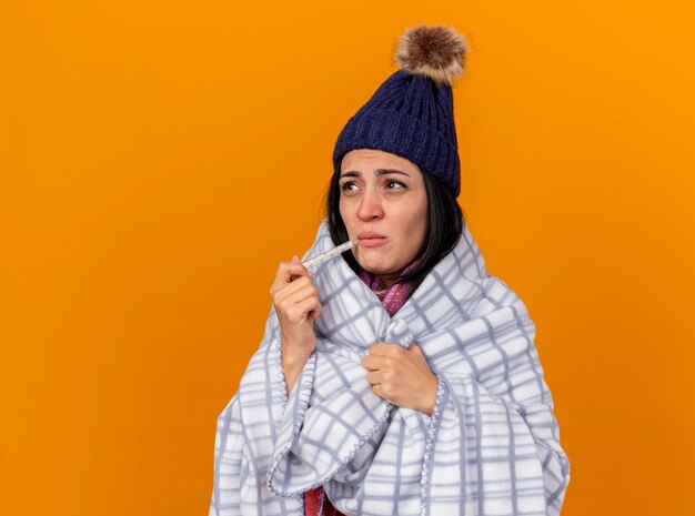 Нахмурившаяся молодая кавказская больная девушка в зимней шапке и шарфе, завернутая в плед, держит во рту термометр, хватает плед, глядя в сторону, изолированную на оранжевом фоне с копией пространства