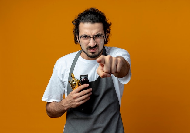 Frowning young caucasian male barber wearing glasses and wavy hair band in uniform holding winner cup and hair clippers and pointing at camera isolated on orange background with copy space