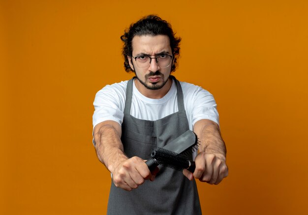 Frowning young caucasian male barber wearing glasses and wavy hair band in uniform stretching out combs towards camera isolated on orange background with copy space