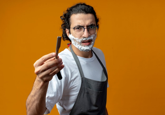 Хмурый молодой кавказский парикмахер в очках и с волнистой лентой для волос в униформе протягивает опасную бритву с кремом для бритья, нанесенным на лицо