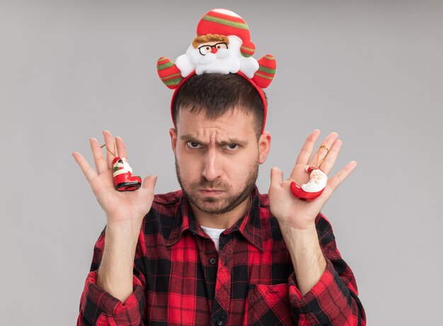 Хмурый молодой кавказский человек в повязке на голову санта-клауса, держащий рождественские украшения санта-клауса, изолированные на белой стене