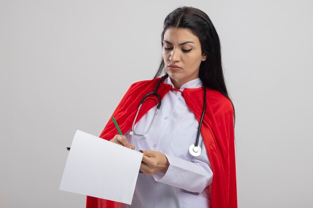 Frowning young caucasian superhero girl wearing stethoscope holding clipboard writing on it with pencil isolated on white background with copy space