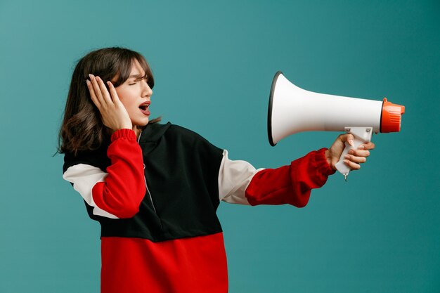 Frowning young caucasian woman stretching speaker out turning head to side pointing speaker at herself closing ear with hand with closed eyes isolated on blue background