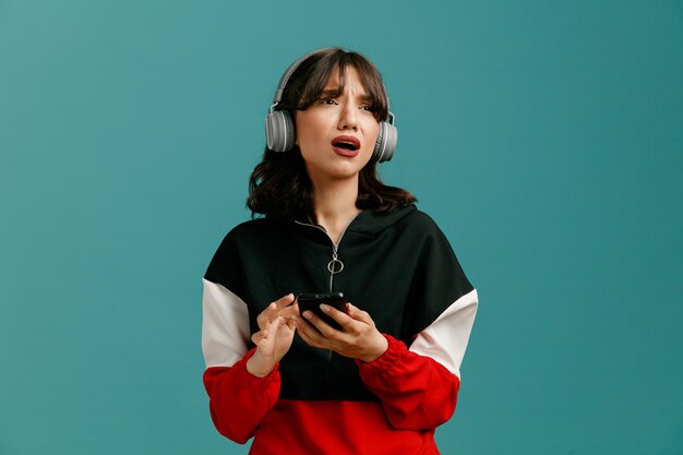 Frowning young caucasian woman wearing headphones holding mobile phone looking at side isolated on blue background