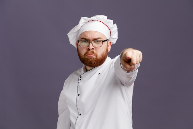 안경 유니폼을 입고 모자를 쓰고 보라색 배경에 격리된 카메라를 보고 가리키는 젊은 요리사
